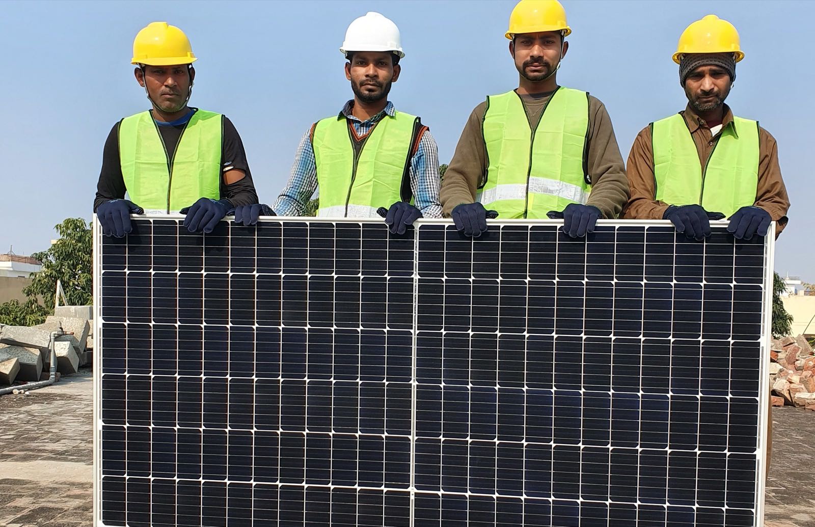 Four installers in hard hats and high-visibility vests standing behind a solar panel on a rooftop in India
